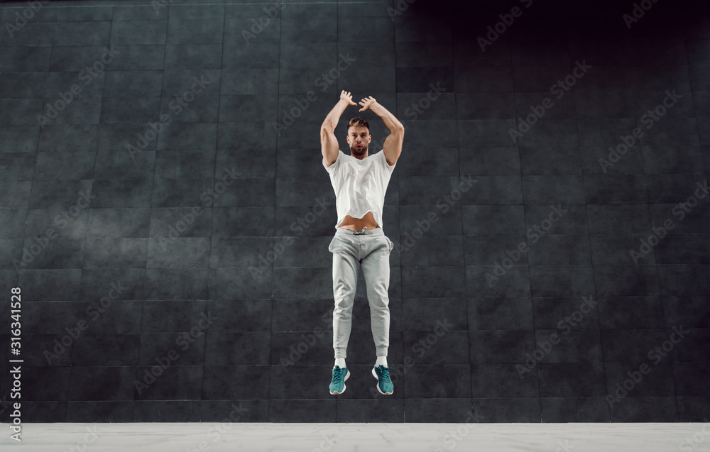 Handsome fit muscular bearded blonde man in tracksuit jumping outside. In background is gray wall.