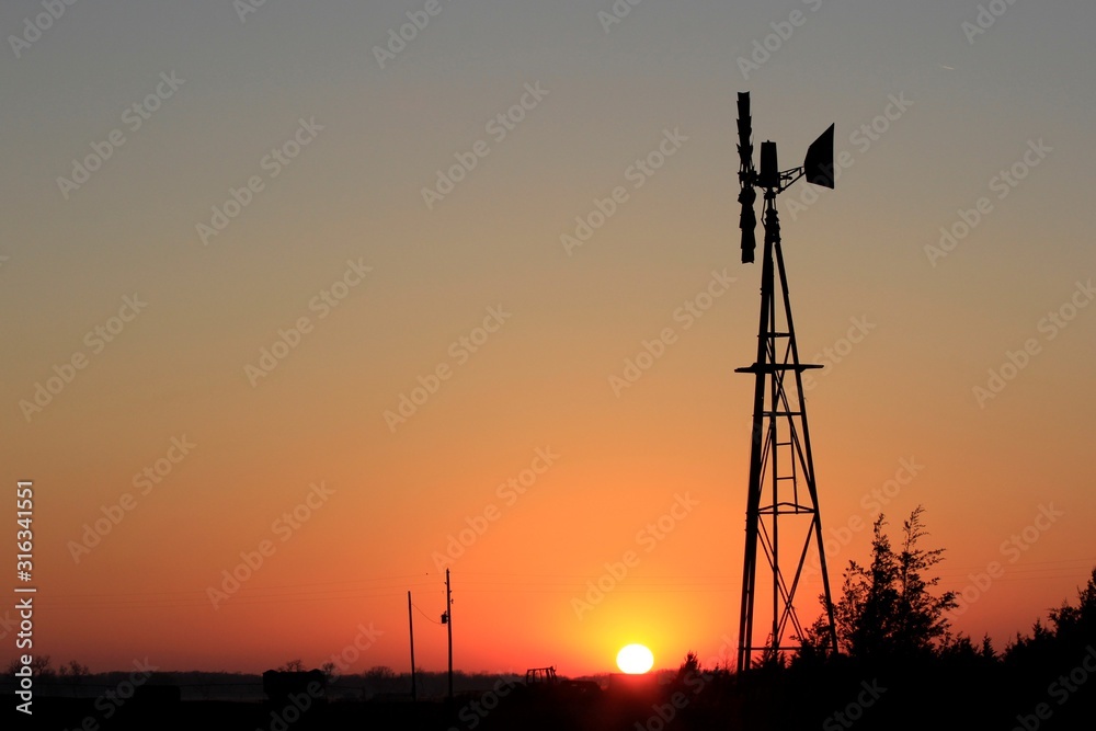 windmill at sunset with powe lines and poles in the background in Kansas.