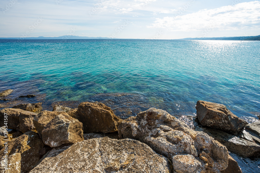 Beautiful sunny summer landscape of blue peaceful sea water, clear sky and big stones in foreground of image. Picturesque Greece nature, Chalkidiki. Horizontal color photography.