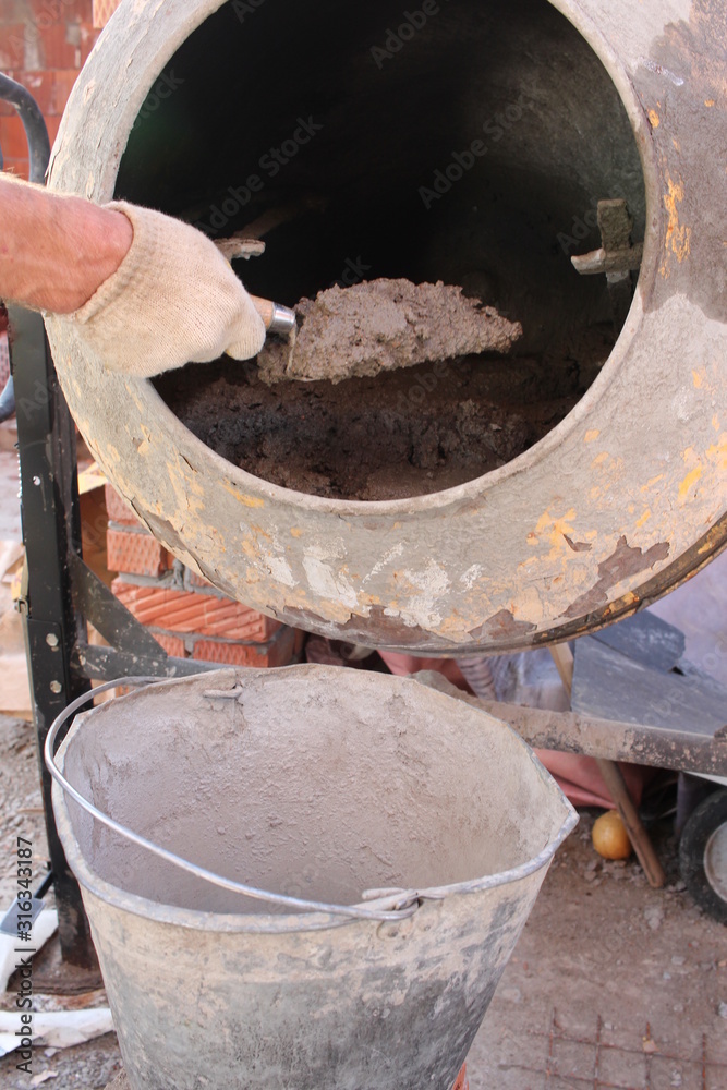 a construction worker puts cement and concrete mortar from a concrete mixer in a bucket at a construction site with his hand a construction worker puts cement and concrete mortar from a concrete mixer in a bucket at a construction site with his hand