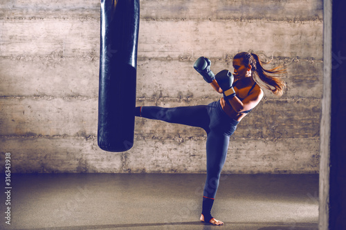 Young fit attractive muscular caucasian female kick boxer in sportswear with boxing gloves kicking boxing bag.
