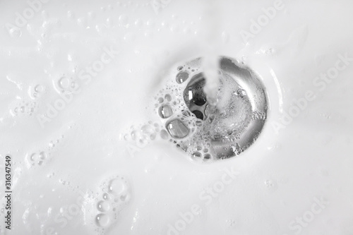 Water with soap foam in white ceramic sink drains down, close up