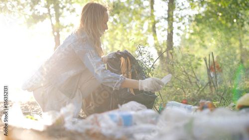 young townswoman is helping to clean territory from waste in forest, collecting garbage in park area in summer day photo