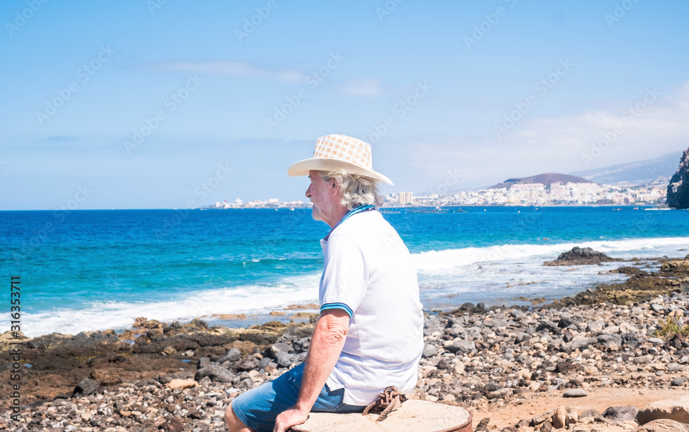 Portrait of a senior man with white hair and beard sitting and looking at the ocean, Blue sky and sea on background. Vacations or retirement. Serenity and relax