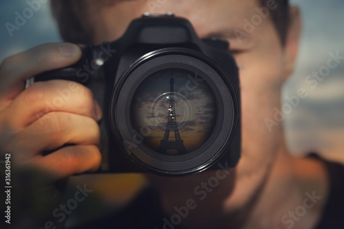 image of a young man with a camera. The lens reflects the Eiffel tower on the background of the sunset. Theme of photography, travel, vacation, trip.