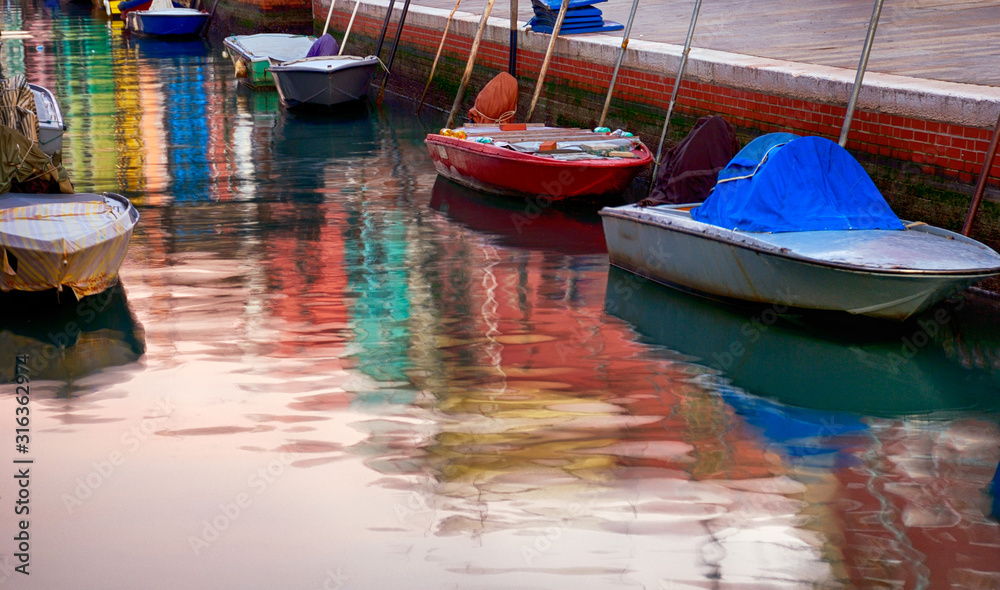 Colorful reflection in canal water of anchored motor boats and painted in vibrant colors houses' facades in Burano, Italy.
