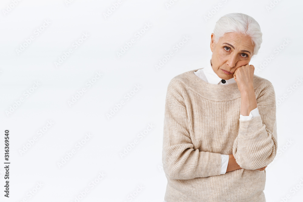 Bored or annoyed senior lady with grey combed hair, lean face on fist, look from under forehead distressed with frustration and bad mood, sulking over white background, insulted or disappointed Bored or annoyed senior lady with grey combed hair, lean face on fist, look from under forehead distressed with frustration and bad mood, sulking over white background, insulted or disappointed