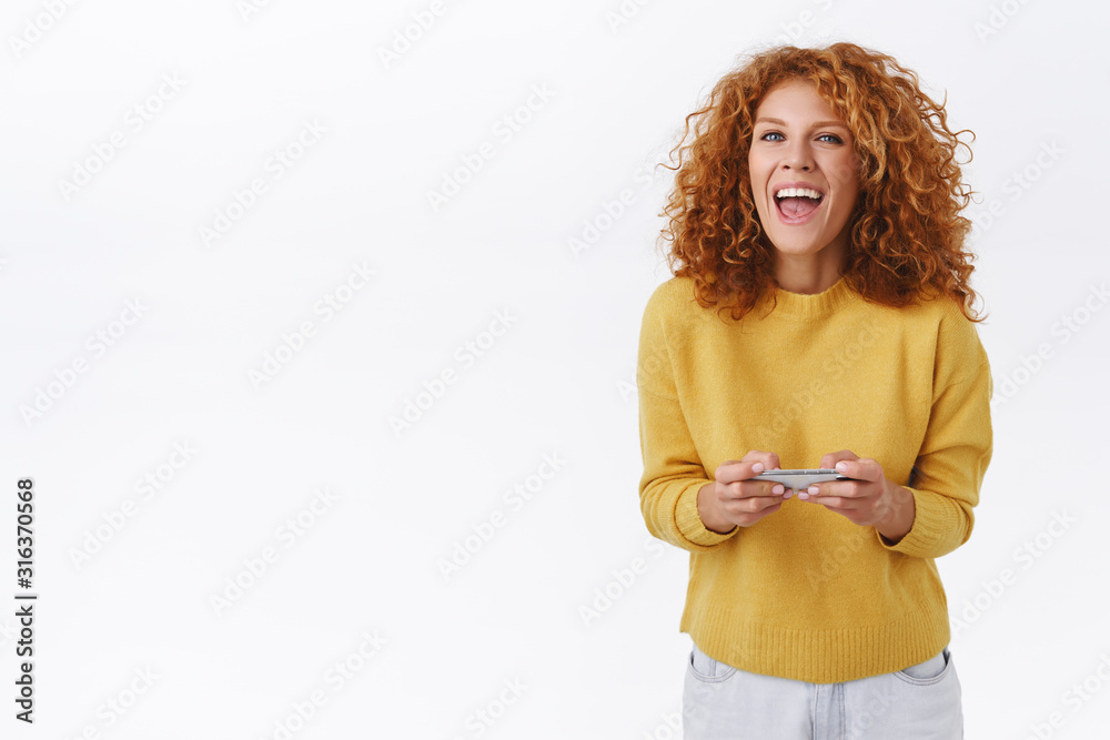Happy, triumphing young attractive redhead woman with curly ginger hair, laughing pleased, holding smartphone horizontally, passed level, beat score and celebrating, become winner, champion