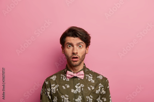 Concerned shocked European man stares with omg expression at camera, gasps from hearing bad terrified news, dressed in fashionable shirt, bowtie stands anxious, isolated over pink background. photo