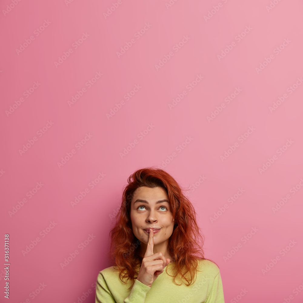 Ginger adult woman presses index finger to lips, prepares surprise, asks not tell anyone, needs stay quiet, wears green jumper, looks upwards, isolated over rosy background. Be quiet, please Ginger adult woman presses index finger to lips, prepares surprise, asks not tell anyone, needs stay quiet, wears green jumper, looks upwards, isolated over rosy background. Be quiet, please