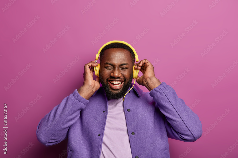 Obraz premium Music fan concept. Smiling black man with dark bristle, uses musical accessory gadget, enjoys perfect sound in headphones, has broad smile, white teeth, closes eyes, dressed in fashionable clothes