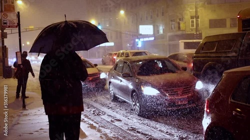 man is standing outside with umbrella in the extreme cold and snow in Chicago area during a blizzard during a winter night in January.  traffic is slowing down due to weather conditions