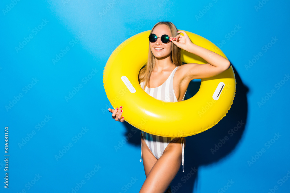 Portrait of beautiful young woman with sunglasses in bikini playing with yellow inflatable ring on blue background