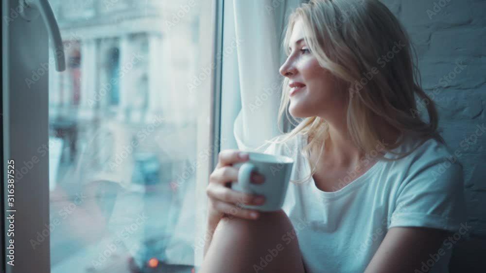 Attractive beautiful young woman sitting on windowsill drinking coffee or tea in modern apartment relax at the morning great view from window cozy home enjoying comfortable relax warm slow motion