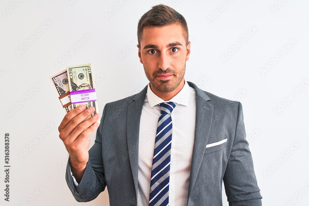 Young handsome business man holding bunch of dollars banknotes over isolated background with a confident expression on smart face thinking serious Young handsome business man holding bunch of dollars banknotes over isolated background with a confident expression on smart face thinking serious