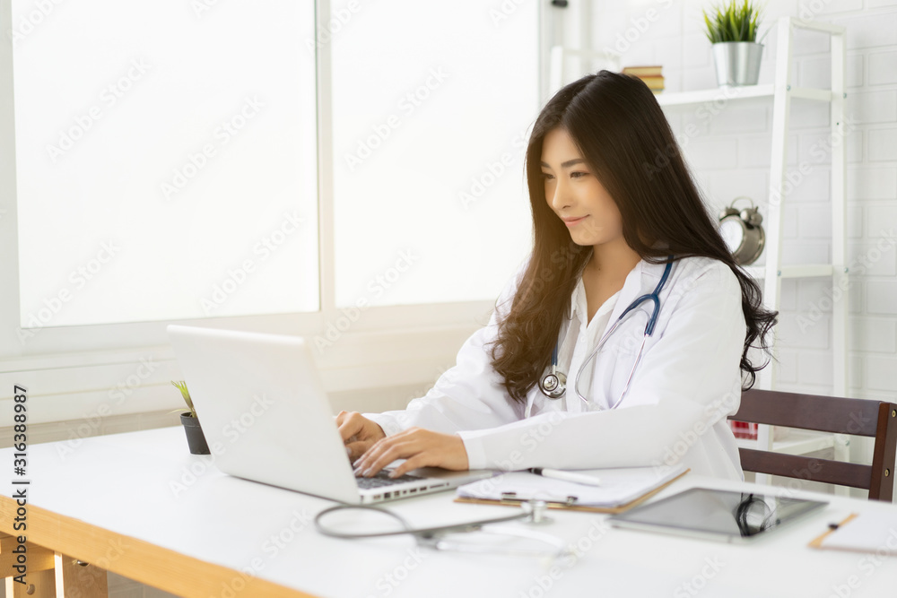 Asian female doctor sitting at hospital office desk giving all patient convenience online service advice and smiling write a prescription on laptop to order medical,health care and preventing disease Asian female doctor sitting at hospital office desk giving all patient convenience online service advice and smiling write a prescription on laptop to order medical,health care and preventing disease