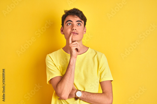 Teenager boy wearing yellow t-shirt over isolated background with hand on chin thinking about question, pensive expression. Smiling with thoughtful face. Doubt concept.