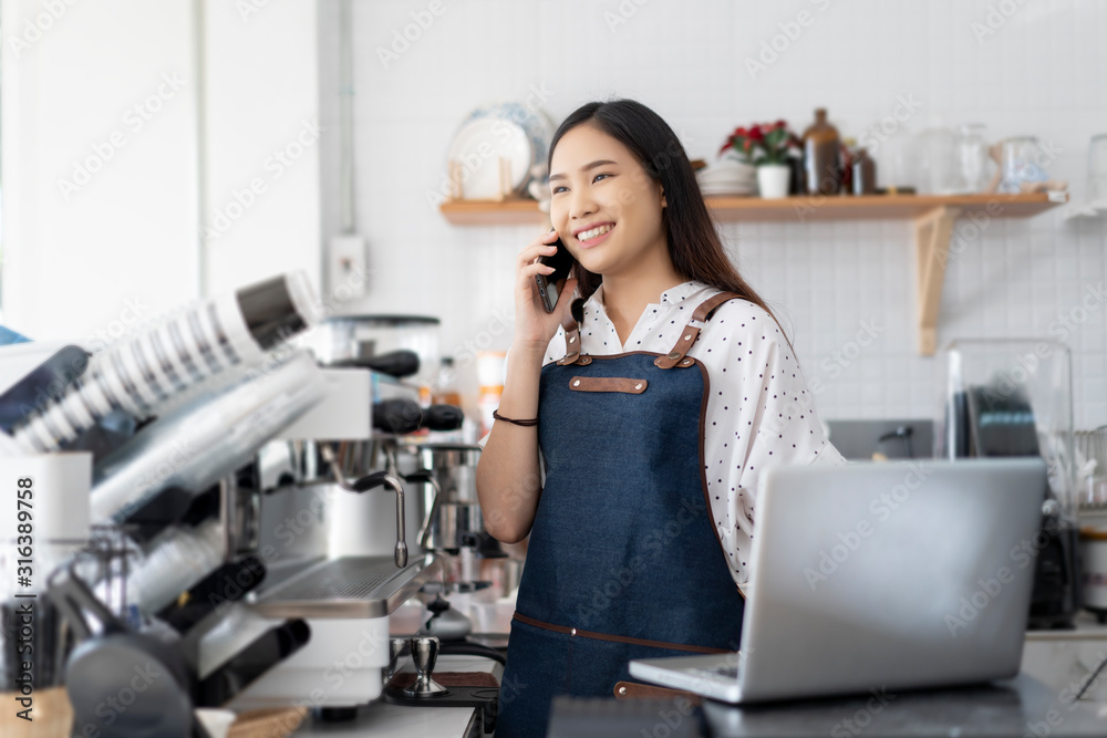 Happy Asian woman barista working on laptop smiling and talking on smartphone, accepting writing order at a coffee shop. waitress in Cafeteria Small business owner, online purchase and service concept Happy Asian woman barista working on laptop smiling and talking on smartphone, accepting writing order at a coffee shop. waitress in Cafeteria Small business owner, online purchase and service concept