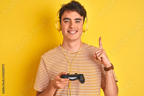 Teenager boy playing video games using gamepad over isolated yellow background surprised with an idea or question pointing finger with happy face, number one