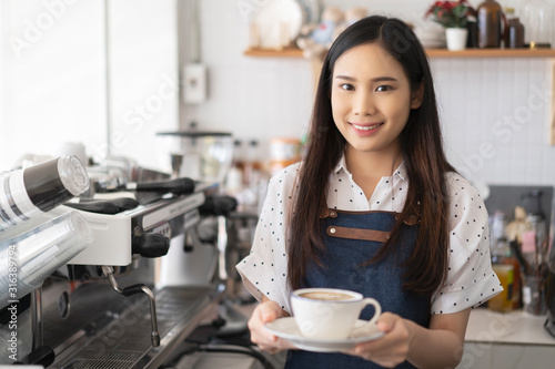 Photography Startup small business (SME) Asian women Smiling Barista waitress holding a white-hot coffee cup of latte with rosetta in a cafe coffee shop serving a client, business owner food and drink concept