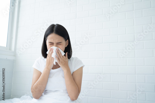 Flu.Rhinitis, cold, sickness, allergy concept. Young Asian woman got nose allergy, flu sneezing nose sitting at bed in bedroom,female. Brunette sneezing in a tissue photo