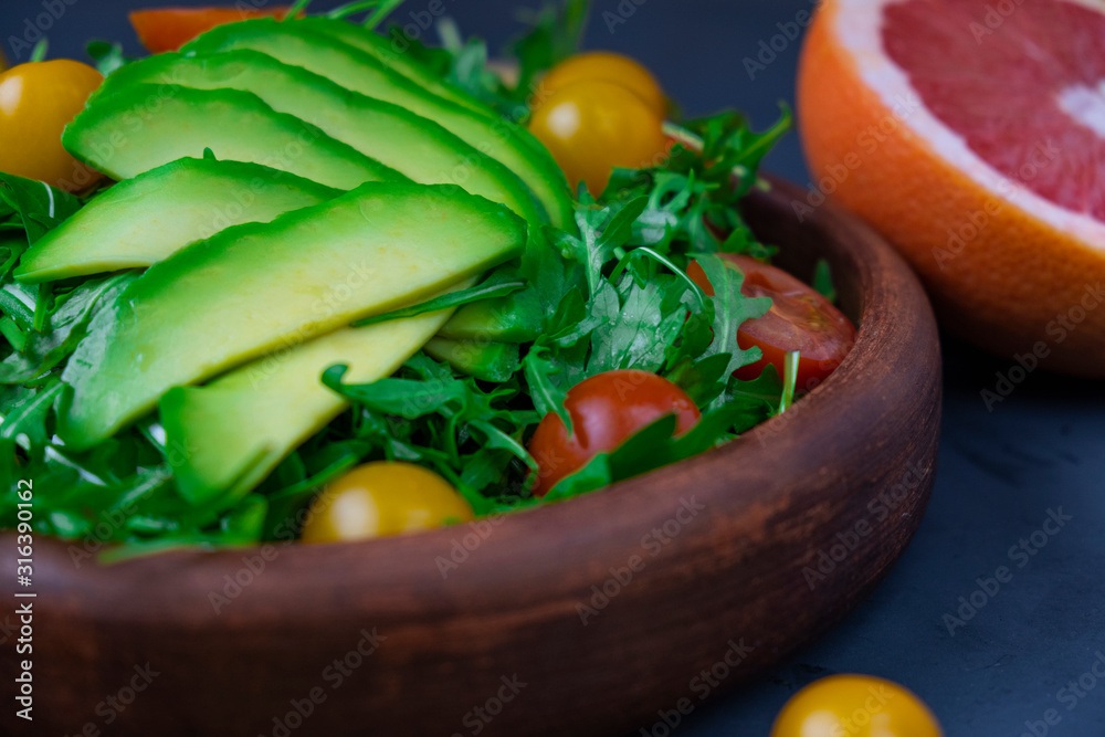 closeup plate with fresh salad with arugula, cherry tomatoes and avocado, bright green on a black background, avacado and grapefruit cut in half
