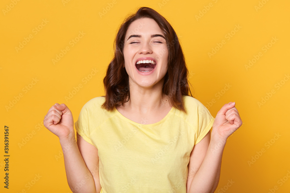 Close up portraiot of happy young woman with beautiful dark hair, clenching fist while looking directly at camera, wearing casual t shirt, isolated over yellow background. People emotions concept. - obrazy, fototapety, plakaty 