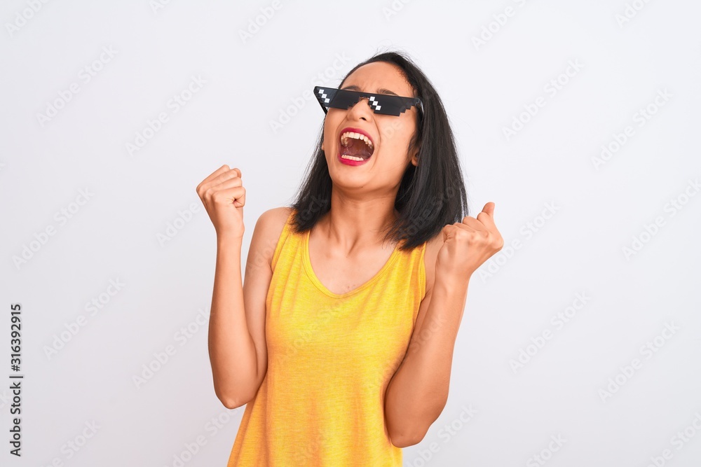 Young beautiful chinese woman wearing thug life sunglasses over isolated white background celebrating mad and crazy for success with arms raised and closed eyes screaming excited. Winner concept Young beautiful chinese woman wearing thug life sunglasses over isolated white background celebrating mad and crazy for success with arms raised and closed eyes screaming excited. Winner concept