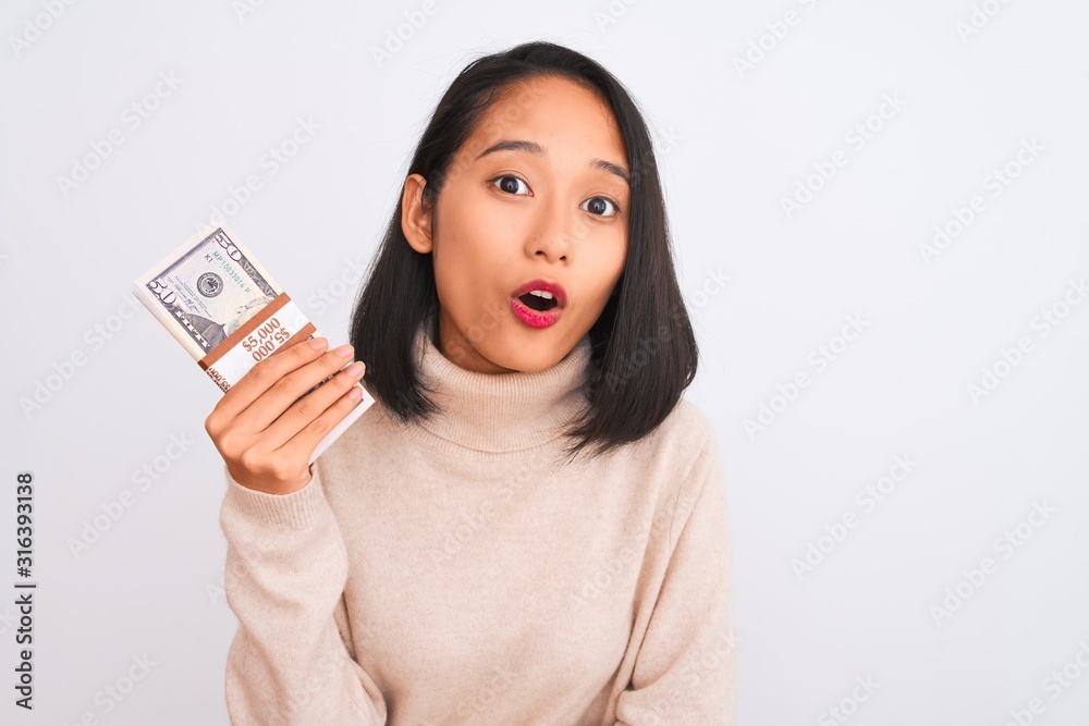 Young beautiful chinese woman holding dollars standing over isolated white background scared in shock with a surprise face, afraid and excited with fear expression Young beautiful chinese woman holding dollars standing over isolated white background scared in shock with a surprise face, afraid and excited with fear expression