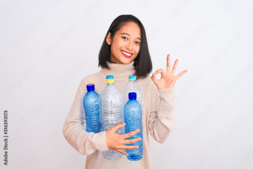 Young beautiful chinese woman recycling plastic bottles over isolated white background doing ok sign with fingers, excellent symbol Young beautiful chinese woman recycling plastic bottles over isolated white background doing ok sign with fingers, excellent symbol