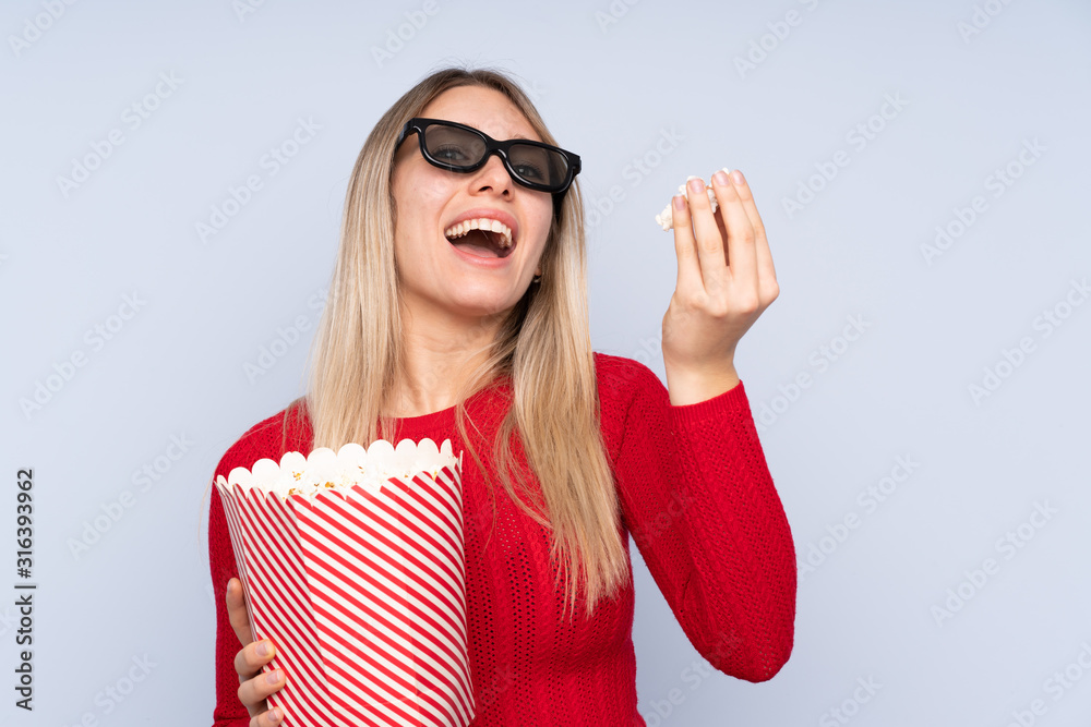 Young blonde woman over isolated blue background with 3d glasses and holding a big bucket of popcorns