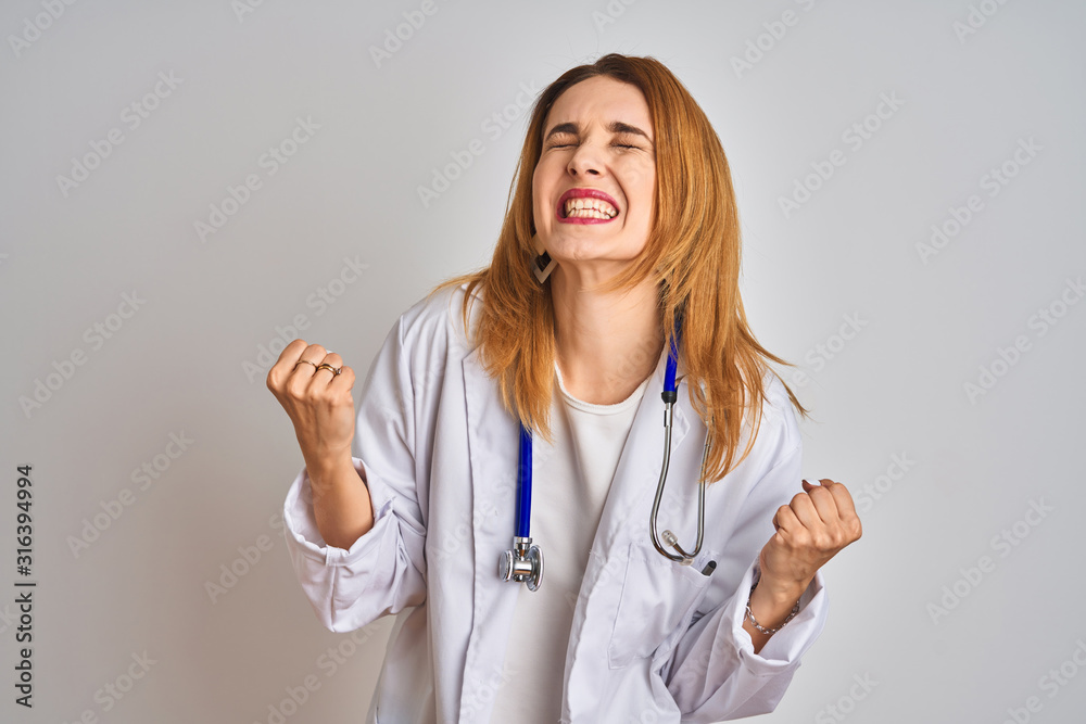 Redhead caucasian doctor woman wearing stethoscope over isolated background very happy and excited doing winner gesture with arms raised, smiling and screaming for success. Celebration concept. Redhead caucasian doctor woman wearing stethoscope over isolated background very happy and excited doing winner gesture with arms raised, smiling and screaming for success. Celebration concept.
