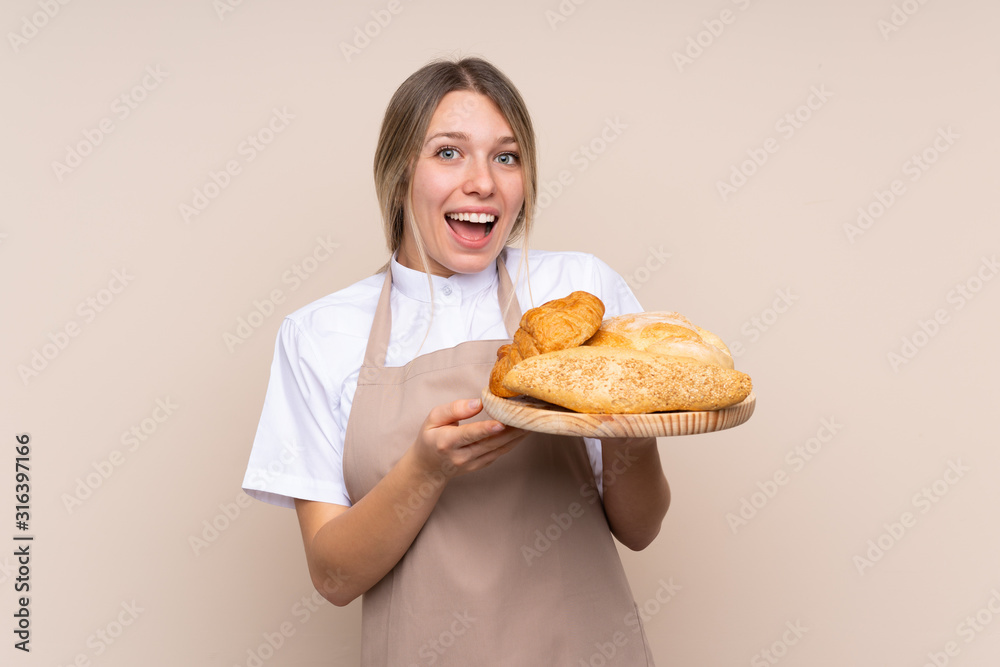Young blonde girl with apron. Female baker holding a table with several breads with surprise and shocked facial expression Young blonde girl with apron. Female baker holding a table with several breads with surprise and shocked facial expression