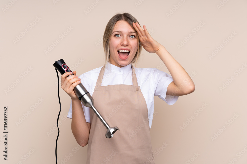 Young blonde woman using hand blender with surprise and shocked facial expression