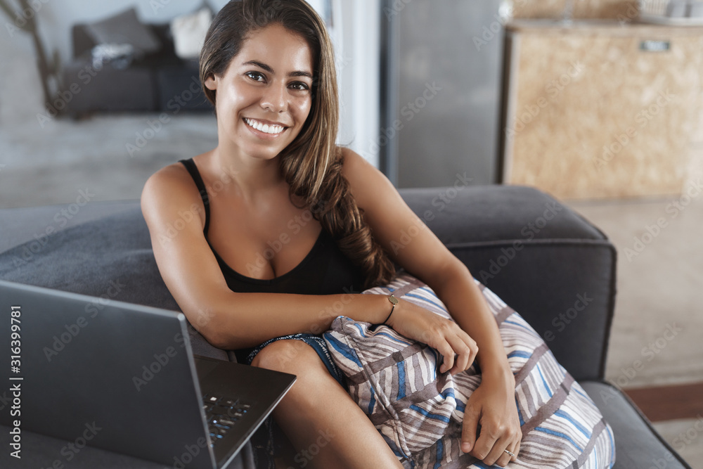Digital nomad, people and business concept. Attractive cheerful young woman sitting on sofa, hugging pillow and smiling camera, watching video laptop, working remote from co-working space