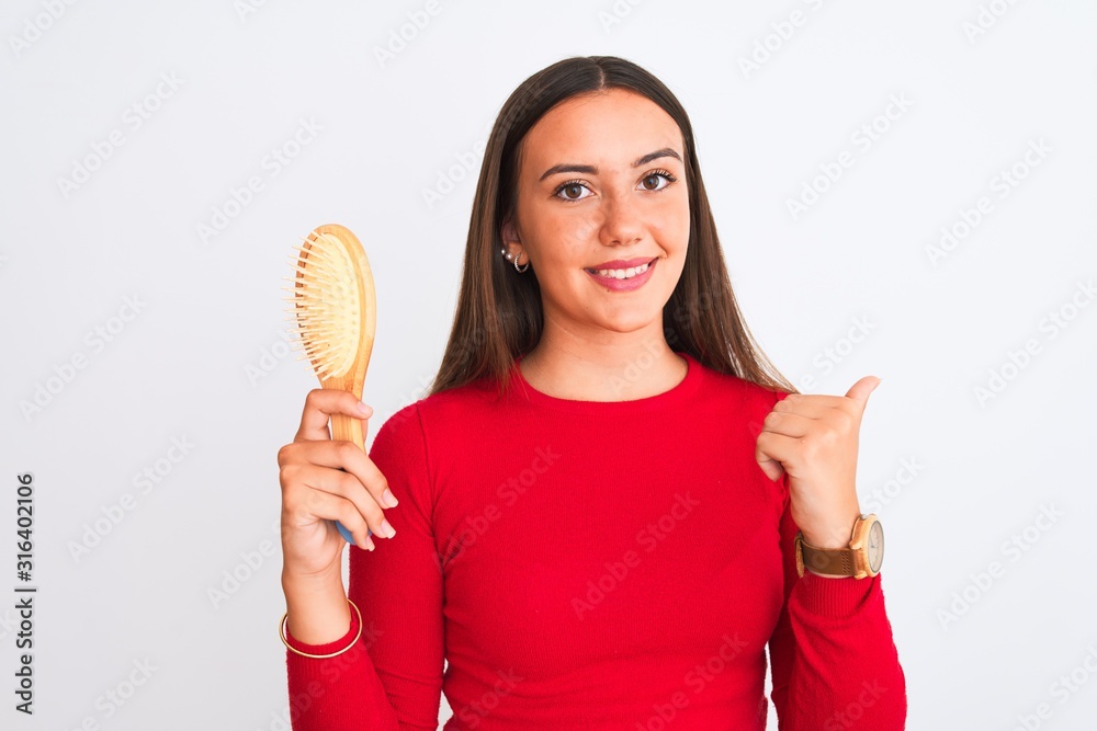 Young beautiful girl holding hair comb standing over isolated white background pointing and showing with thumb up to the side with happy face smiling Young beautiful girl holding hair comb standing over isolated white background pointing and showing with thumb up to the side with happy face smiling
