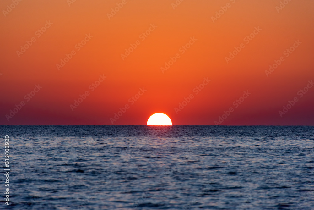 Sunset on the sea. The sun disk plunges below the water on the horizon at the end of the day creating a beautiful fiery red sky over the calm blue sea
