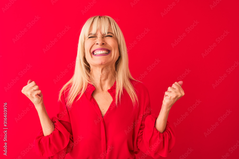 Middle age woman wearing elegant shirt standing over isolated red background very happy and excited doing winner gesture with arms raised, smiling and screaming for success. Celebration concept. Middle age woman wearing elegant shirt standing over isolated red background very happy and excited doing winner gesture with arms raised, smiling and screaming for success. Celebration concept.