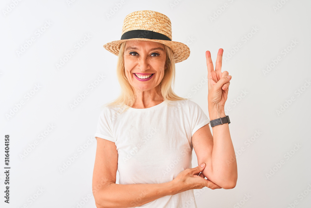 Middle age woman wearing casual t-shirt and hat standing over isolated white background smiling with happy face winking at the camera doing victory sign. Number two.