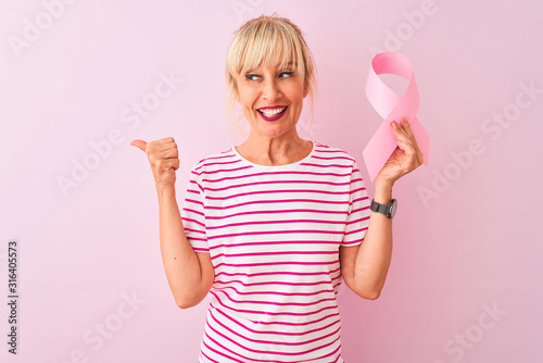 Middle age woman holding cancer ribbon standing over isolated pink background pointing and showing with thumb up to the side with happy face smiling