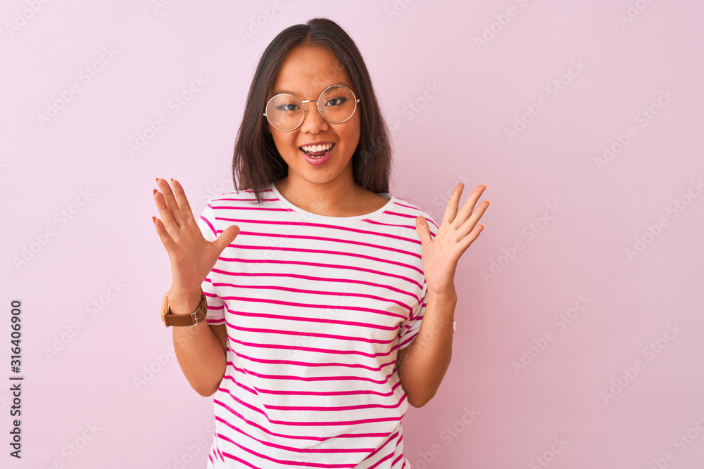 Young chinese woman wearing striped t-shirt and glasses over isolated pink background celebrating crazy and amazed for success with arms raised and open eyes screaming excited. Winner concept Young chinese woman wearing striped t-shirt and glasses over isolated pink background celebrating crazy and amazed for success with arms raised and open eyes screaming excited. Winner concept