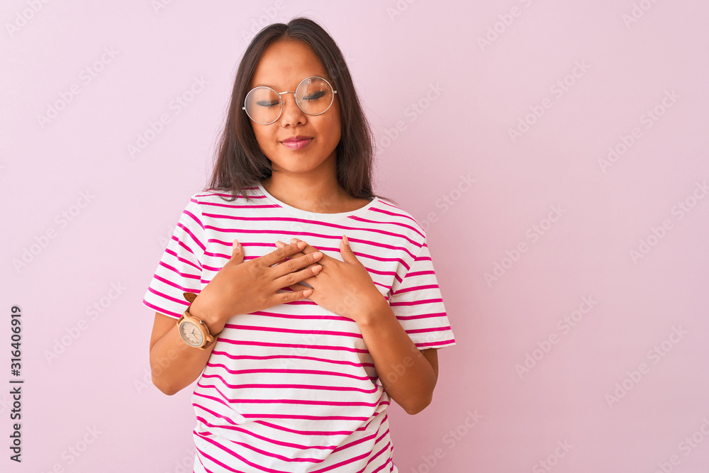 Young chinese woman wearing striped t-shirt and glasses over isolated pink background smiling with hands on chest with closed eyes and grateful gesture on face. Health concept. Young chinese woman wearing striped t-shirt and glasses over isolated pink background smiling with hands on chest with closed eyes and grateful gesture on face. Health concept.