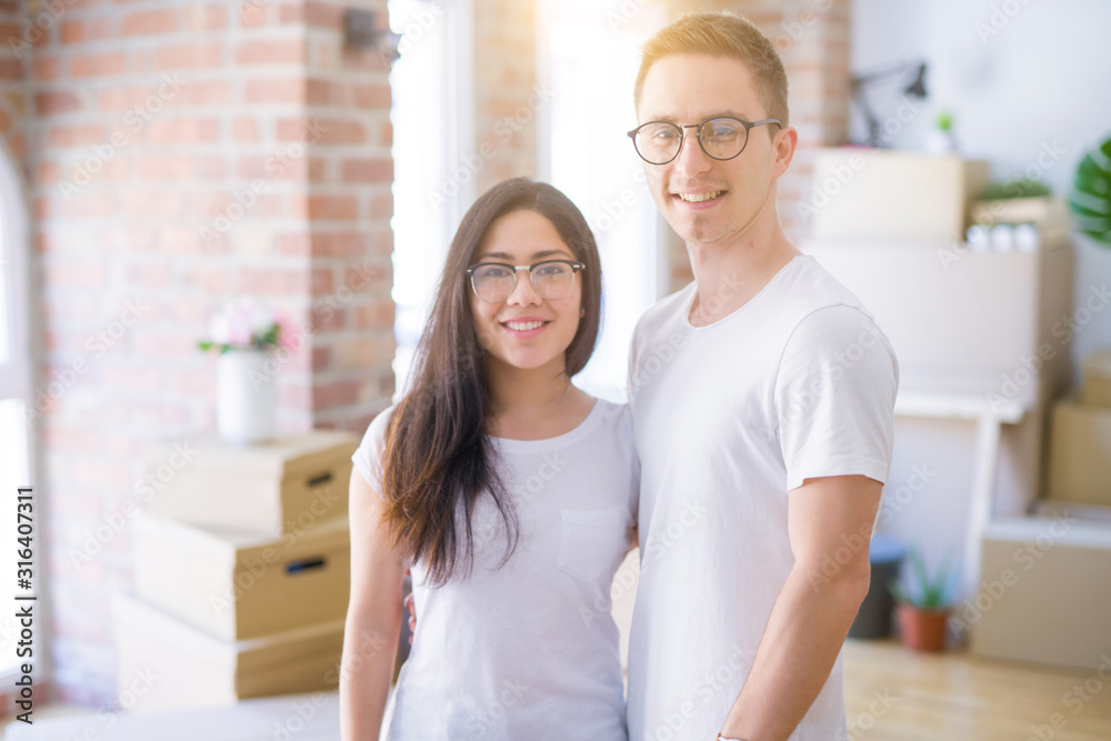 Young beautiful couple wearing glasses standing at new home around cardboard boxes
