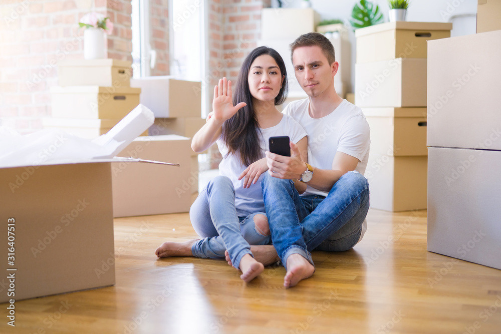 Beautiful couple sitting on the floor using smartphone at new home around cardboard boxes with open hand doing stop sign with serious and confident expression, defense gesture Beautiful couple sitting on the floor using smartphone at new home around cardboard boxes with open hand doing stop sign with serious and confident expression, defense gesture