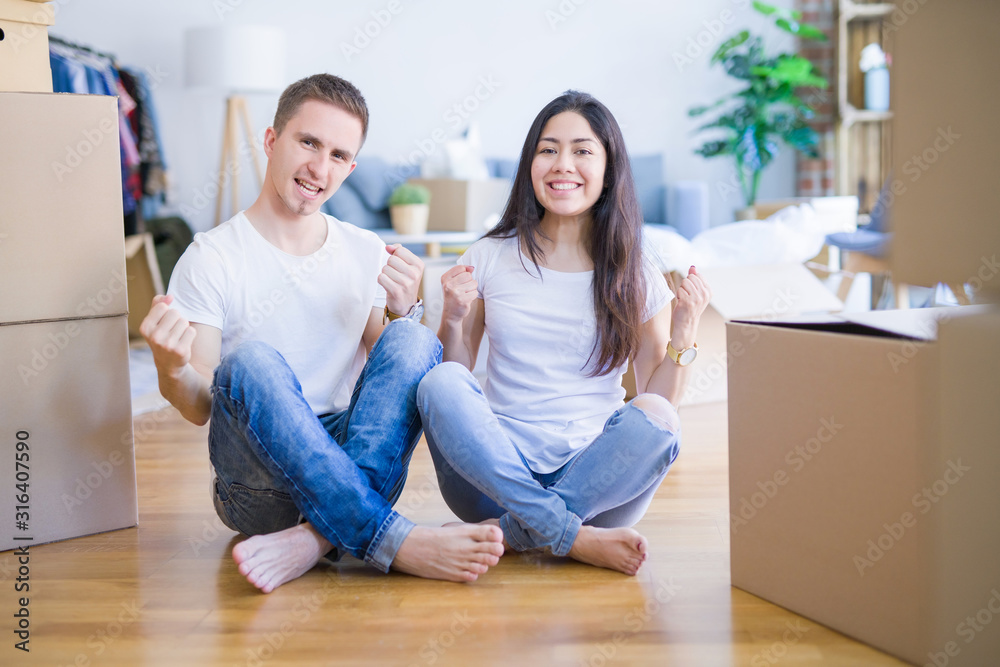 Young beautiful couple sitting on the floor at new home around cardboard boxes celebrating surprised and amazed for success with arms raised and open eyes. Winner concept. Young beautiful couple sitting on the floor at new home around cardboard boxes celebrating surprised and amazed for success with arms raised and open eyes. Winner concept.