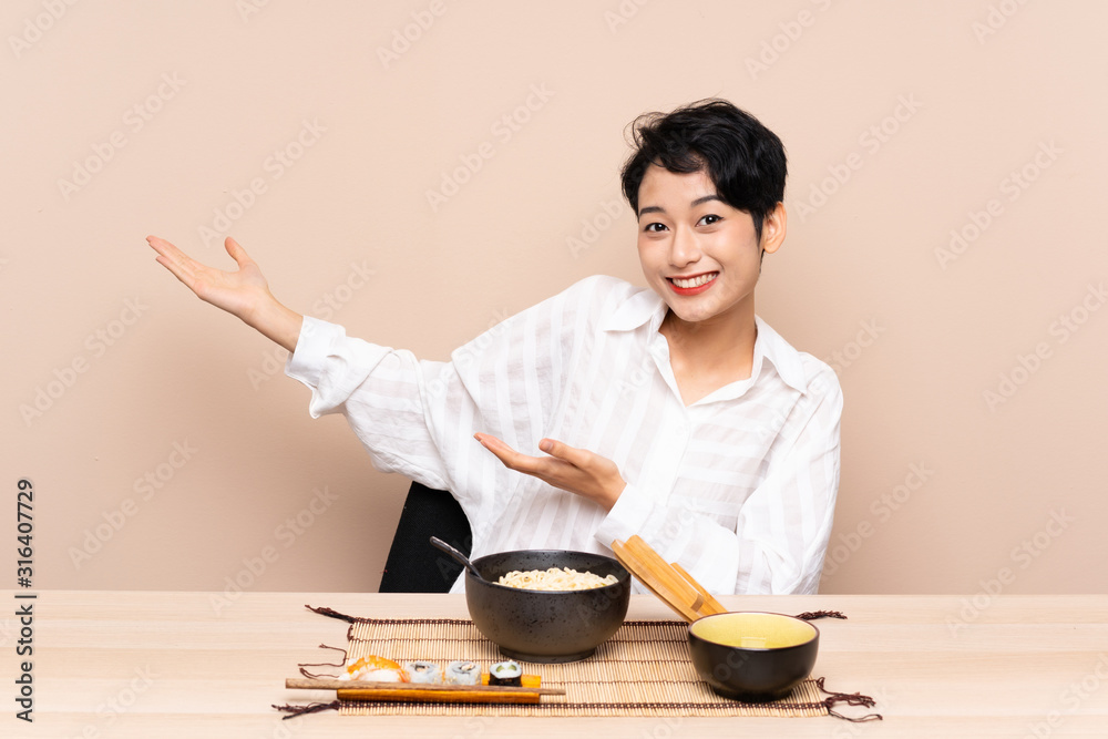 Young Asian girl in a table with bowl of noodles and sushi extending hands to the side for inviting to come