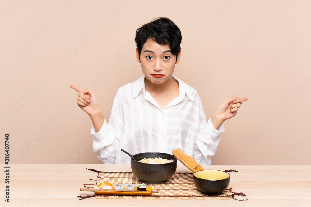 Young Asian girl in a table with bowl of noodles and sushi pointing to the laterals having doubts Young Asian girl in a table with bowl of noodles and sushi pointing to the laterals having doubts