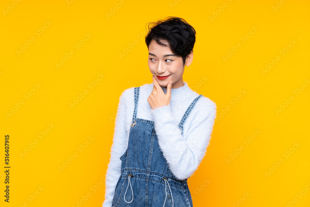 Young Asian girl in overalls over isolated yellow background thinking an idea Young Asian girl in overalls over isolated yellow background thinking an idea