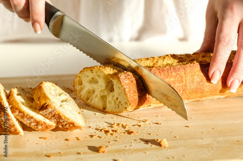 Slicing tasty, fresh, crisp bread. Beautiful female hands, a knife for cutting bread and sliced baguette closeup. Floury product on a cutting board. Healthy eating concept.