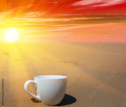closeup porcelain coffee cup on the sand at the dramatic sunset, stylized coffee background
